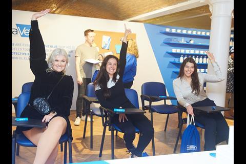 Three customers sat at school desks and raising one hand at CeraVe pop-up, Covent Garden